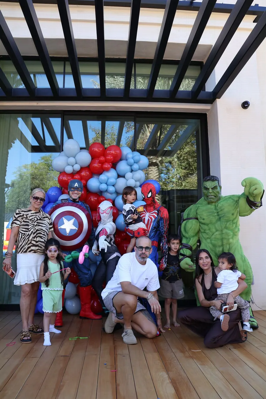 Family and kids posing with superhero characters in front of a red and blue balloon wall at a party, photographed by Lihi Matzri