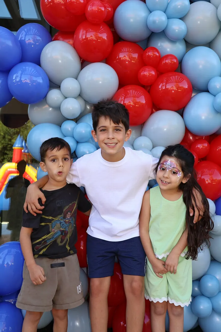 Three smiling kids standing together in front of a red and blue balloon wall at a party, photographed by Lihi Matzri